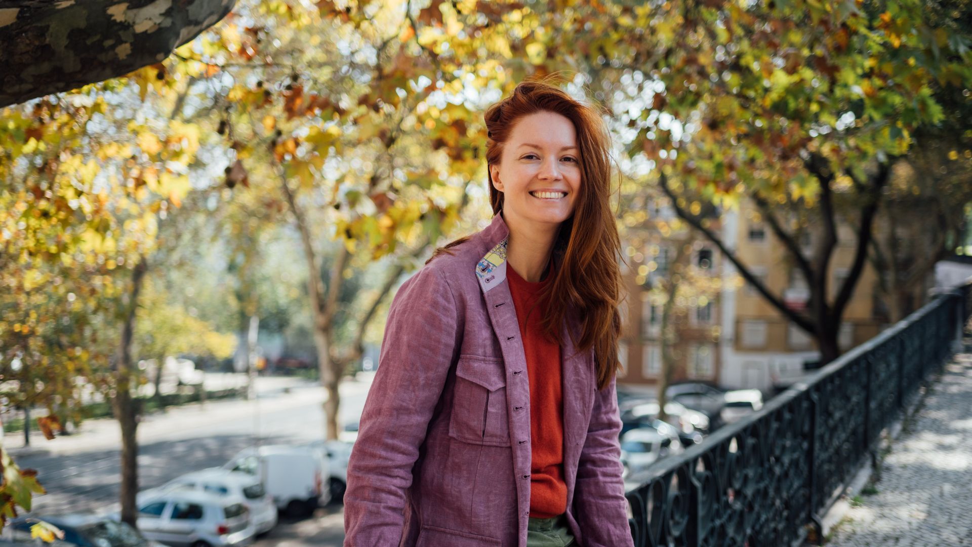 Eine Frau mir roten, langen Haaren lehnt sich an ein Geländer und lächelt fröhlich in die Kamera.