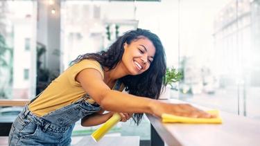 Eine junge Frau im Overall lächelt, während sie in einem hell erleuchteten Raum mit großen Fenstern eine Oberfläche mit einem gelben Tuch reinigt.