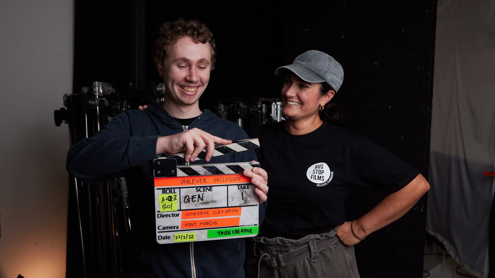 Ein Mann hält eine Filmklappe. Eine Frau mit Schirmmütze steht daneben und lächelt ihn an.